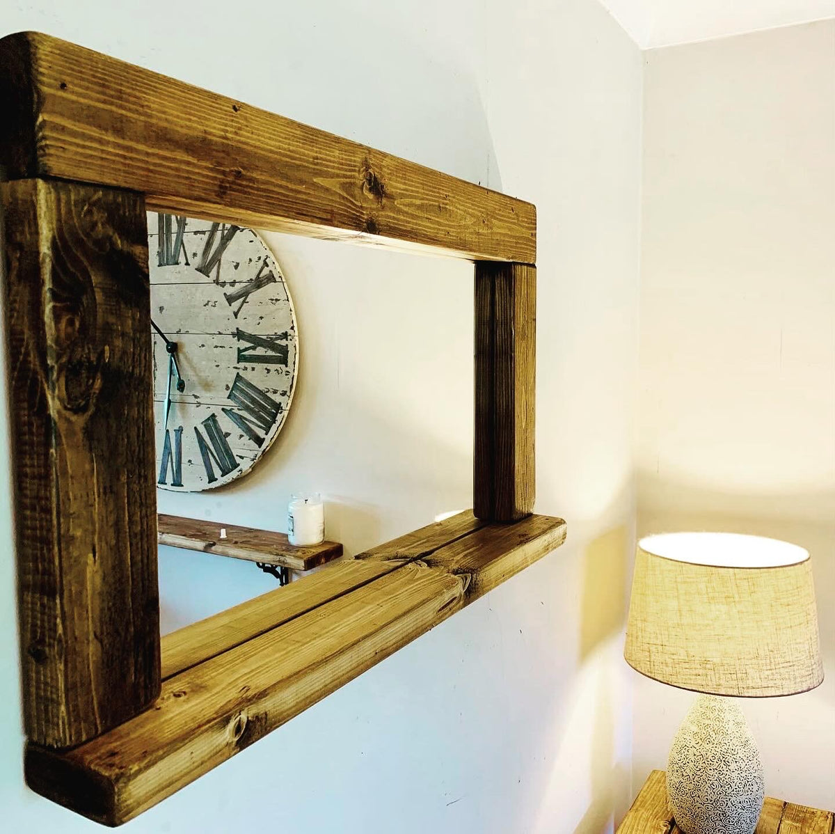A rustic rectangular wooden mirror with a thick frame and shelf, mounted on a white wall in a room setting with a clock and lamp visible in the background.