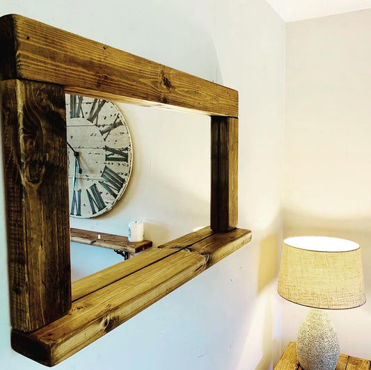 A rustic rectangular wooden mirror with a thick frame and shelf, mounted on a white wall in a room setting with a clock and lamp visible in the background.